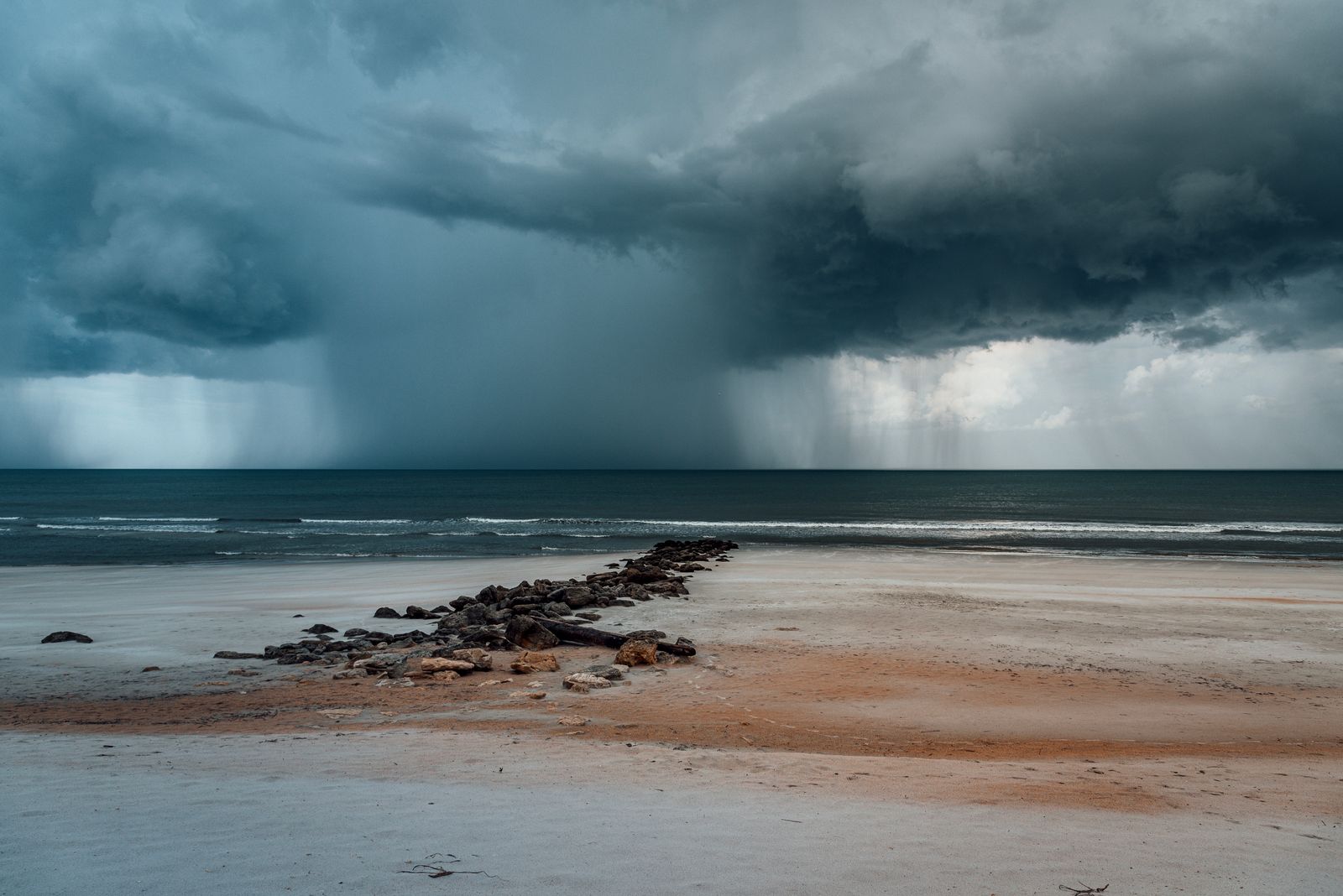 Dark storm clouds dropping heavy rain over the ocean as a squall moves toward a sandy beach and rock jetty