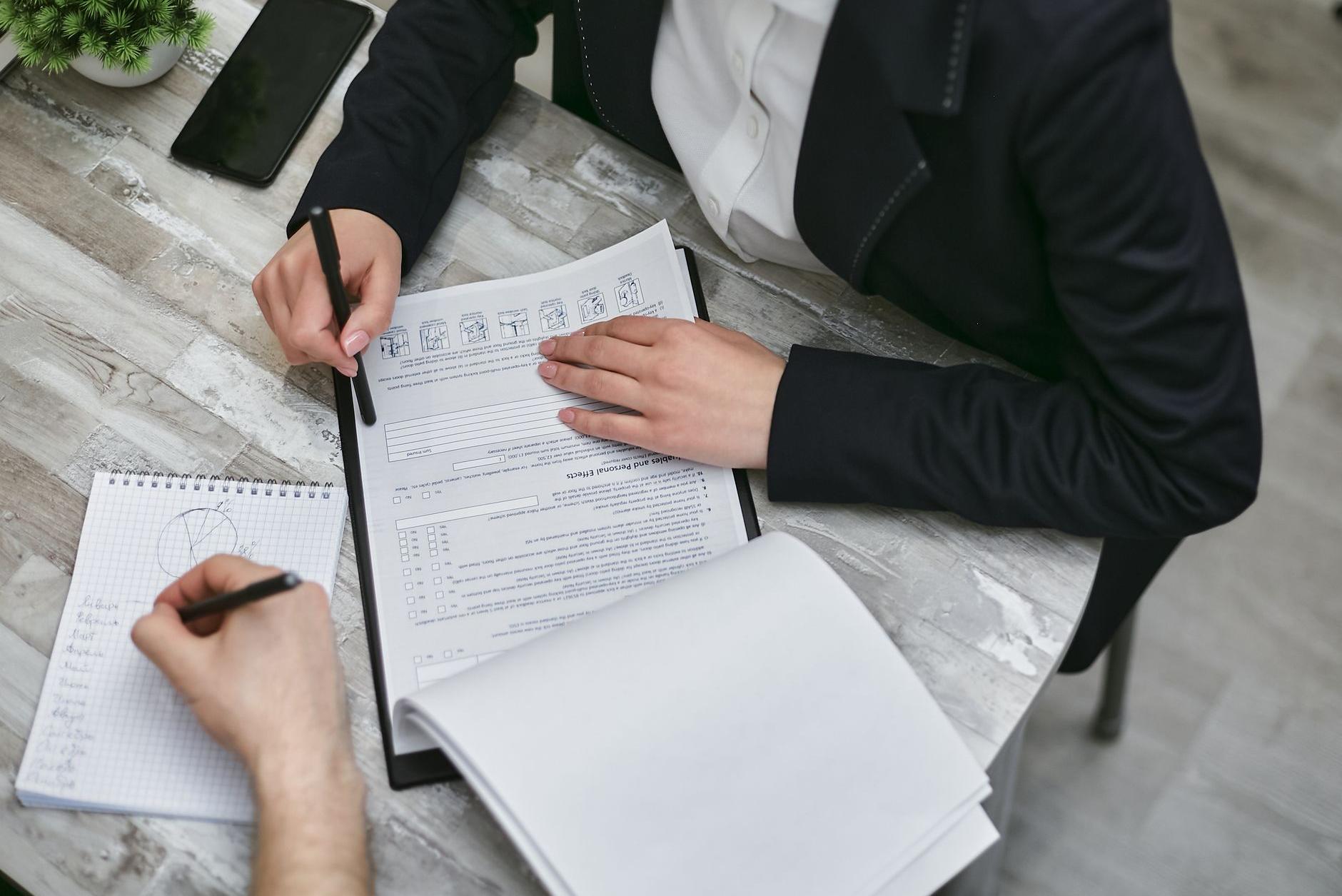 Two people at a table with a paper form and a notepad, one holding a pen over the form