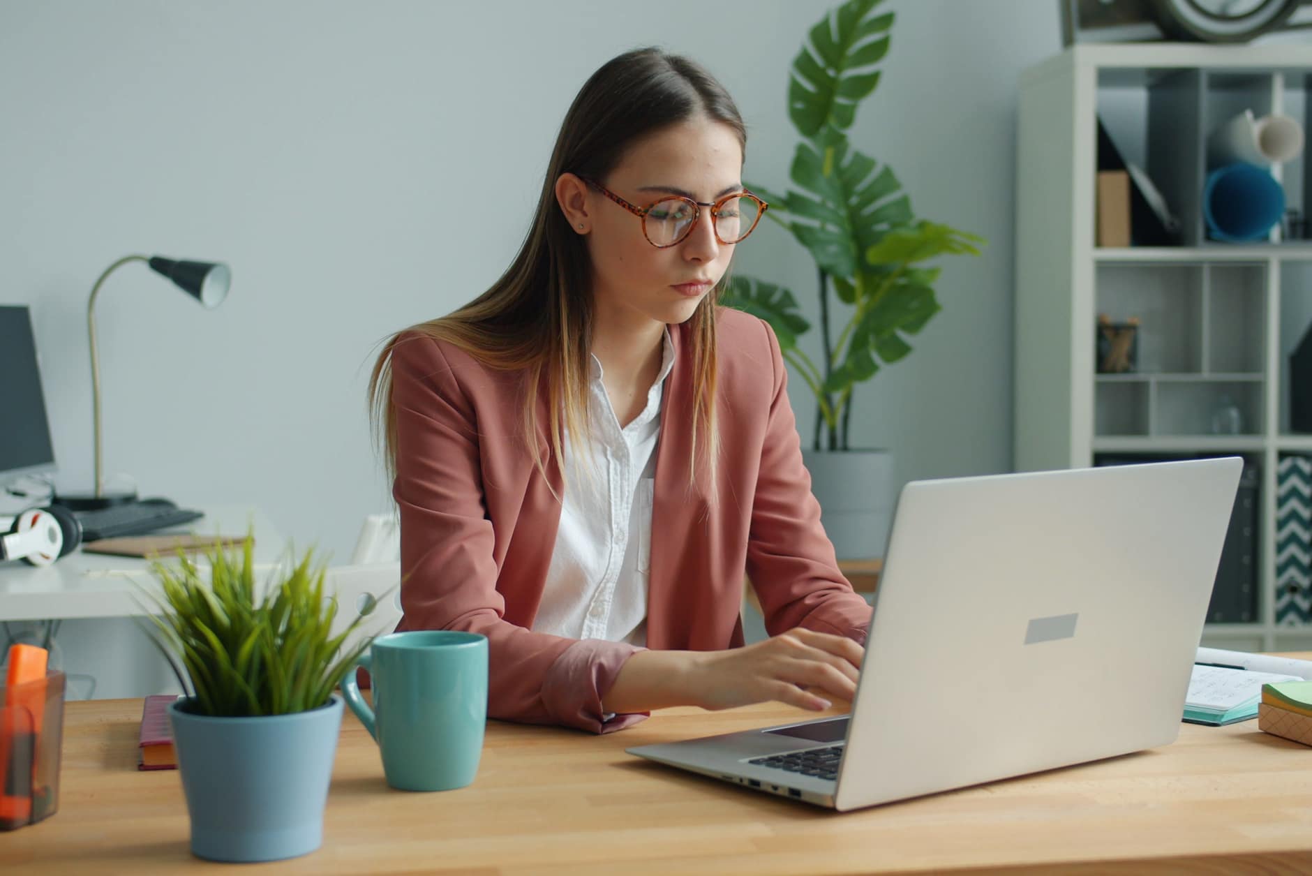 Woman in glasses and a pink blazer typing on a laptop at a wooden desk in a small office